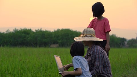 Farmer mother and daughter looking rice field and checking data in laptop on sunset background, happy Asian people family working and playing in rice farmland of rural or countryside, education family