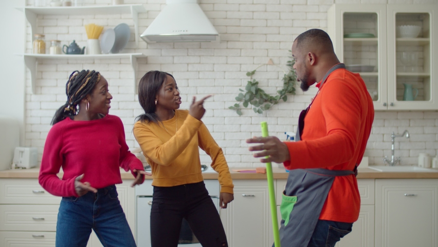 Portrait of cheerful handsome african american single father with mop and two adorable teenage girls taking a break, having fun dancing in domestic kitchen while making housekeeping together.