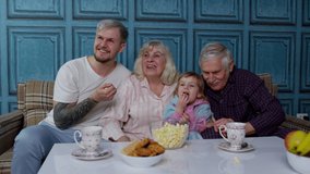 Multigenerational happy family laughing, watching comedy cartoons television movies eating popcorn at home. Senior couple grandfather and grandmother relaxing with adult son and girl kid granddaughter