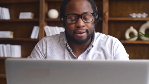 Smiling young African-American office worker in glasses and headset looking at the laptop webcam and talking, positive black man on video call working in the customer service department, front view