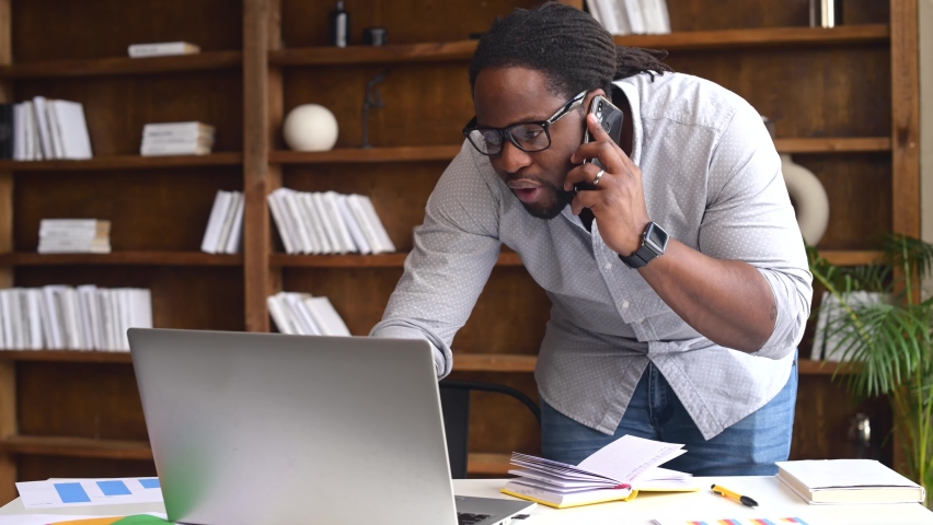 Busy young African-American male office worker wearing glasses standing near the desk, has a phone call and typing on laptop, multitasking guy working on a project in a modern office | Shutterstock HD Video #1068983557