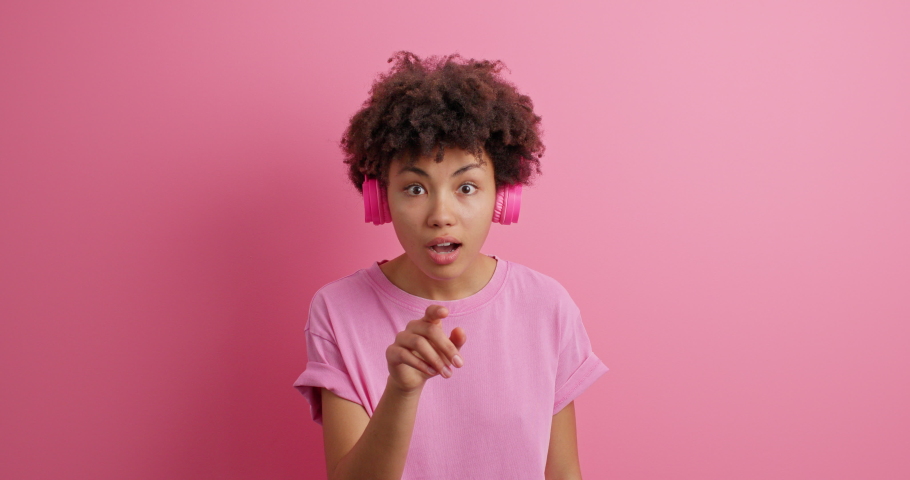 Wow how icredible it is. Impressed curly haired young woman indicates in front says wow wears casual t shirt wireless headphones on ears notices something amazing isolated over pink background | Shutterstock HD Video #1068984631