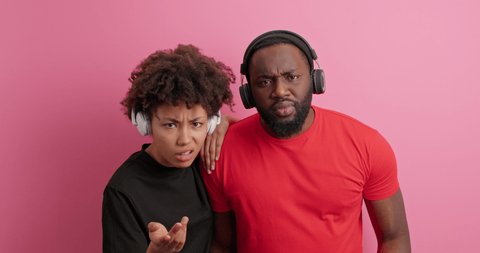 Puzzled displeased ethnic woman and man with dark skin look indignant at each other shrug shoulders listen music viw wireless headphones cannot understand why something happened so pose indoor