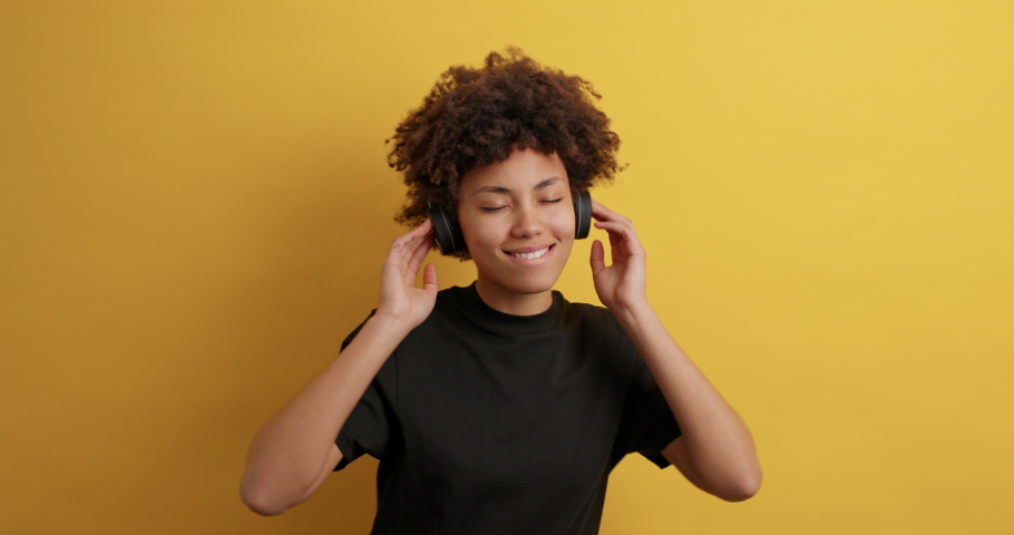 Happy energetic pretty African American woman enjoys playlist composition wears wireless headphones with good sound quality dances carefree dressed in black t shirt isolated on yellow studio wall | Shutterstock HD Video #1068984700
