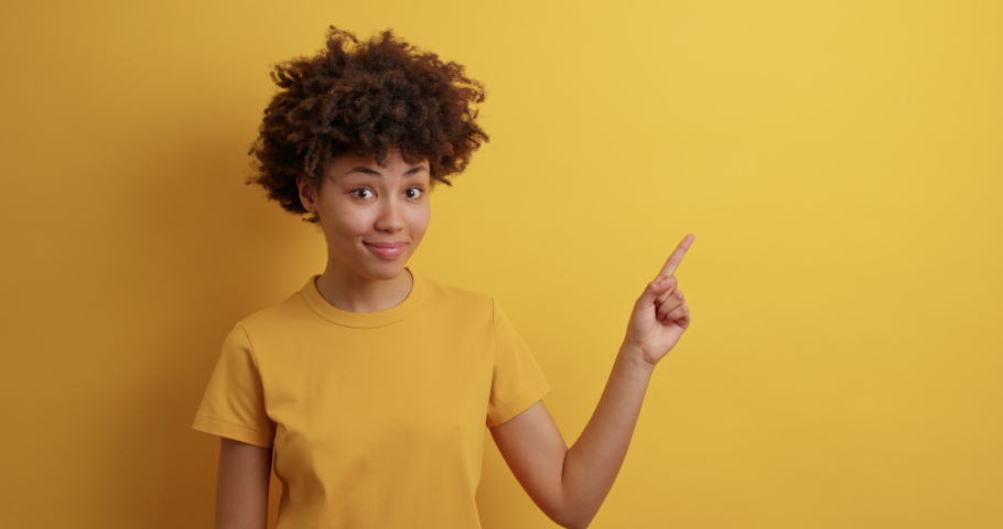 Positive curly haired woman smiles positively recommends using blank space indicates at upper right corner dressed in casual t shirt isolated over vivid yellow background shows direction or way | Shutterstock HD Video #1068984742