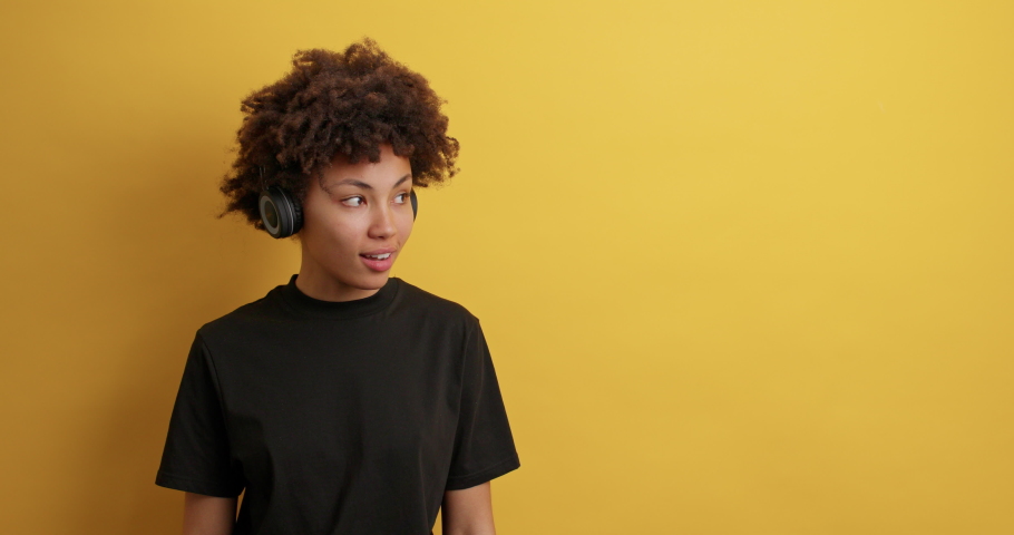 Glad dark skinned teenage girl talks about promo offer asks to take look and check out banner smiles happily listens music via wireless headphones wears black t shirt isolated over yellow background | Shutterstock HD Video #1068984757