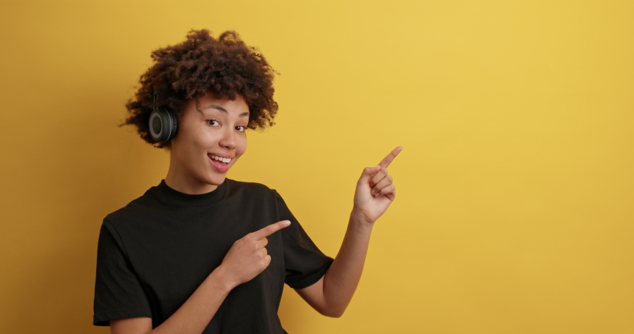 Smiling African American woman shows good deal offer indicates at copy space with good mood listens music via headphones shows advertisement or logo banner makes announcement dressed casually | Shutterstock HD Video #1068984769