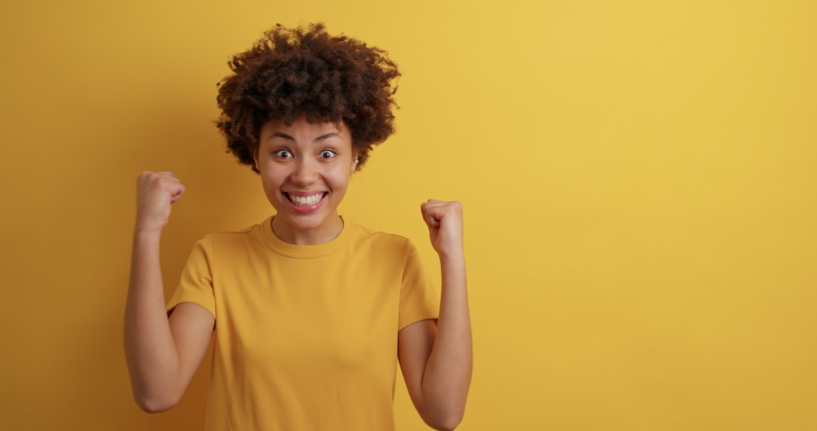 Yes I am winner. Positive Afro American woman clenches fists achieves goal feels like champion dressed in casual clothes isolated over yellow background with copy space area glad to recieve prize Royalty-Free Stock Footage #1068984817