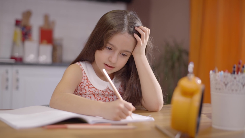 Smart little schoolgirl sitting at the desk at home. Happy cute girl child elementary school student learning at the table, doing homework, writing in the exercise book. | Shutterstock HD Video #1068987604