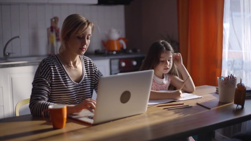 In a dim light in the evening, the little girl is doing her classes, chatting and laughing while her mother is working on the computer in the kitchen. Royalty-Free Stock Footage #1068987607