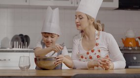 Cute little son and his mother are learning to cook together. Mother and son wearing chef hats are having fun together while trying to bake a cake in the kitchen. Slow motion video.