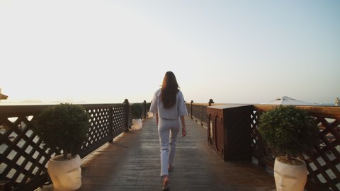 Low angle view of woman walking on wooden pier during sunset. Back view of attractive female tourist walking on pier with Dubai cityscape in the background