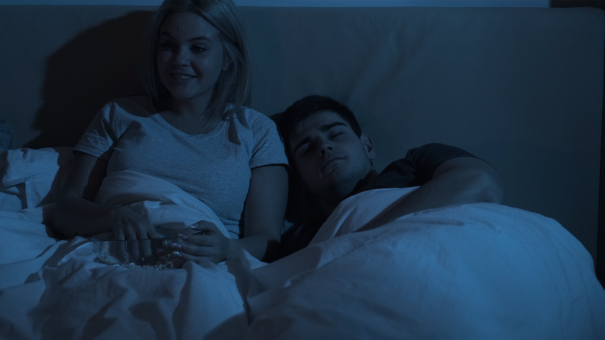Night movie. Late entertainment. Home leisure. Excited curious woman enjoying watching fun TV show eating popcorn in bed with tired man sleeping in dark bedroom with blue flicker light. | Shutterstock HD Video #1068996166
