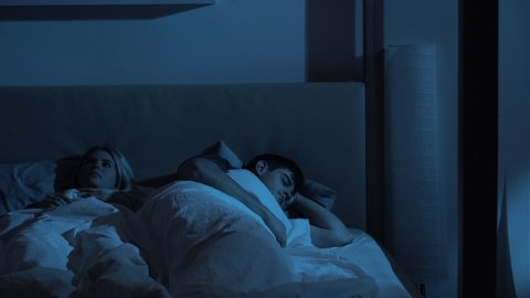 Night insomnia. Couple issue. Bedtime problem. Exhausted woman annoyed with loud noise punching snoring husband sleeping in bed in dark blue light.