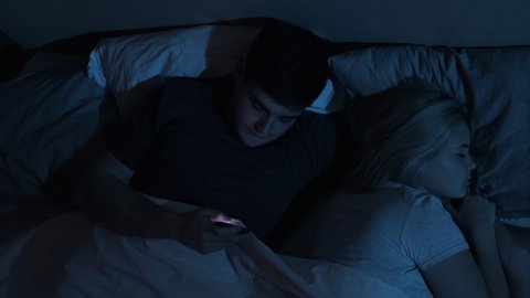 Cheating husband. Couple issue. Infidelity betrayal. Disloyal man using mobile phone for chatting to lover lying in bed with sleeping wife late at night in dark blue light.