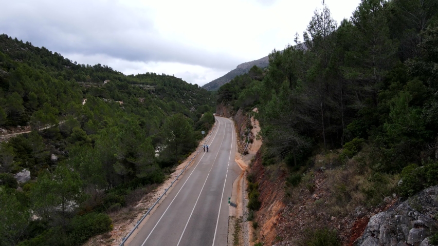 Panning drone footage of three team cyclists riding along a picturesque Spanish road, through a valley with exposed red earth, a river gorge and lush green trees lining the road