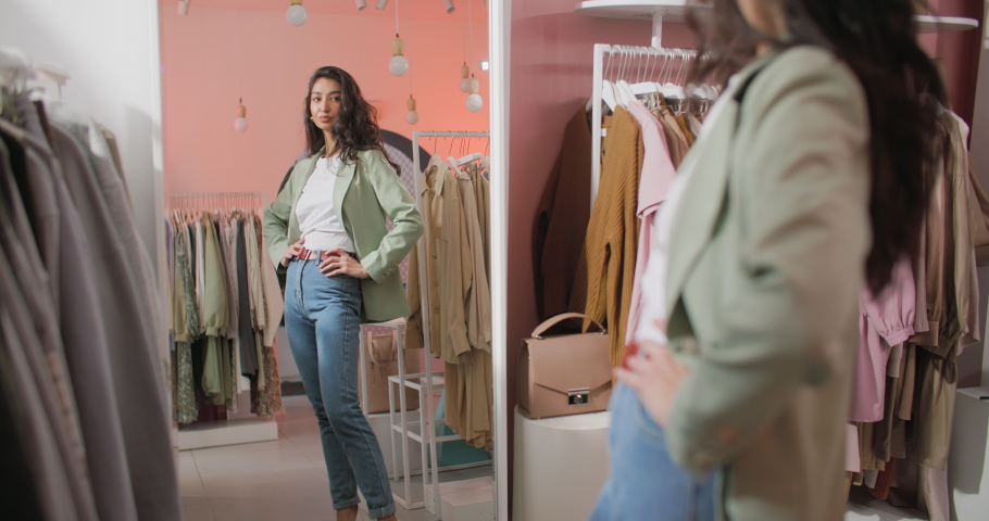 A Back View Shooting of a Brunette Woman Buyer Being in a Clothing Shop Standing in Front of The Mirror and Trying on a Jacket. Beautiful Cheerful Young Woman Choosing Clothes Royalty-Free Stock Footage #1069009477