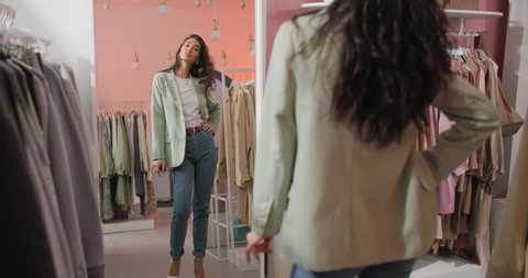 A Back View Shooting of a Brunette Woman Buyer Being in a Clothing Shop Standing in Front of The Mirror and Trying on a Jacket. Beautiful Cheerful Young Woman Choosing Clothes