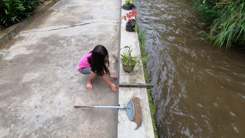 Pretty adorable beautiful cute asian little girl wearing pink shirt enjoy playing broom using hands with joy happy fun on edge of dirty brown small river near street house neighborhood in daylight.