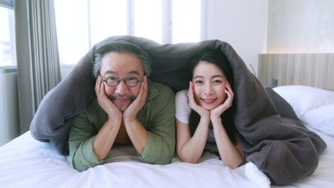 Happiness and joyful mature adult Asian marry couple family mom dad laying laugh smile on white soft bed in bedroom relax casual leisure weekend activity, top view handheld shot slowmotion