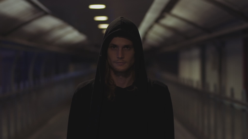 Half-body portrait shot of a young caucasian man with full beard, black hood over his head as he walks towards the camera with a dangerous lookin his eyes.