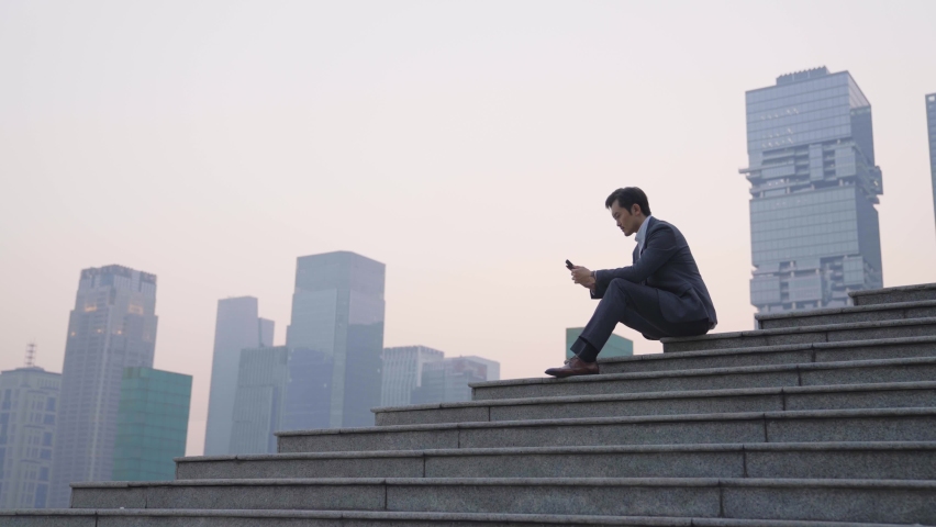 Asian business man sitting on top of steps using cellphone and thinking with modern building background | Shutterstock HD Video #1069015102