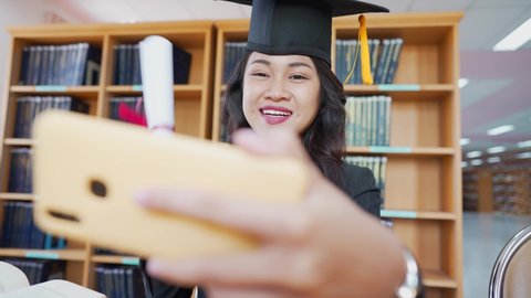 Smiling female Asian student is celebrating her graduation in a term of social distancing by video conference on a smartphone.