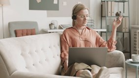 Blond-haired Caucasian girl reclining with legs on couch with laptop on top of them communicating, drinking from ceramic cup. Young woman wearing headset having video conference at home