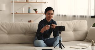 Happy attractive millennial indian female influencer sitting on sofa in front of smartphone camera, recording video at home. Smiling pleasant skilled multiracial woman filming self-presentation.