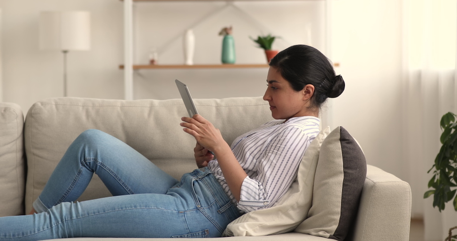 Relaxed young indian biracial woman lying on comfortable couch, involved in web surfing information on digital tablet, communicating in social network, using dating application on weekend at home. Royalty-Free Stock Footage #1069025485
