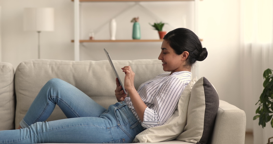 Relaxed young indian biracial woman lying on comfortable couch, involved in web surfing information on digital tablet, communicating in social network, using dating application on weekend at home. | Shutterstock HD Video #1069025485