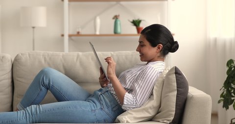 Relaxed young indian biracial woman lying on comfortable couch, involved in web surfing information on digital tablet, communicating in social network, using dating application on weekend at home.