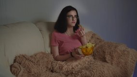 brunette pretty girl with glasses eating potato chips and watching tv at home.