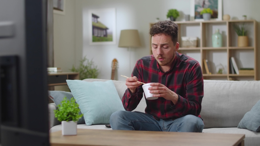 Young handsome man lunchs Chinese noodle from delivery and watching tv at home in the living room.