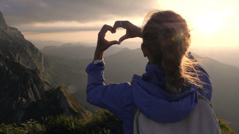 Woman on a hike on top of mountain makes heart shaped finger frame with her hands 