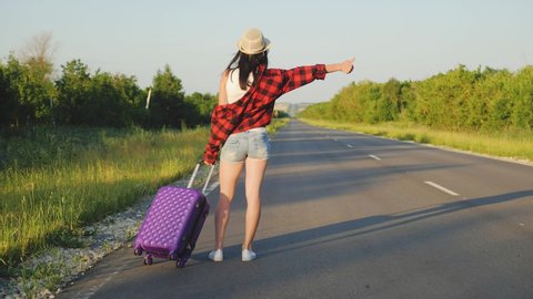The concept of traveling and hitchhiking. A young female traveler hitchhiking and stopping car with thumbs up on road. A female hitchhiker by the road during vacation trip. Summer vacation adventure.