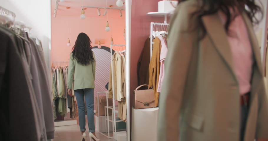 A Back View Shooting of a Brunette Woman Buyer Being in a Clothing Shop Standing in Front of The Mirror and Trying on a Jacket. Beautiful Cheerful Young Woman Choosing Clothes Royalty-Free Stock Footage #1069043671