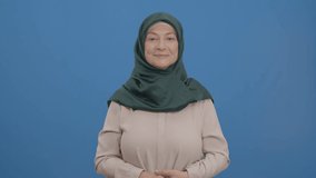The old woman in a turban looks at the advertising space at the bottom of the screen and points with two fingers. Indoor studio shot isolated on blue background. Slow motion video.