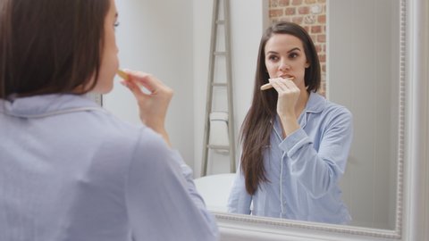 Woman wearing pyjamas walks into modern bathroom and looks into bathroom mirror before squeezing toothpaste onto brush and brushing teeth