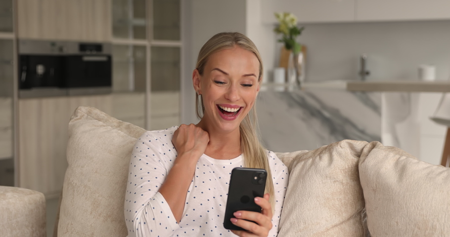 Young beautiful blond woman sit on couch at home hold smart phone read super news feeling excitement celebrate success. Winner and lottery victory moment, sale and discounts, special offer get concept Royalty-Free Stock Footage #1069058761