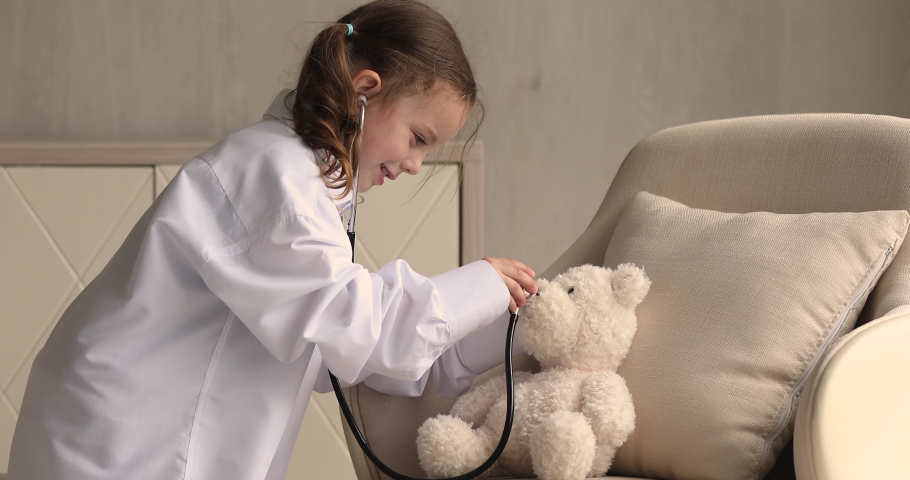 Little girl wear white coat play doctor with plush bear sit on chair. Caring child hold stethoscope touch toy have fun pretending nurse treating fluffy patient at home. Paediatrics, playtime concept Royalty-Free Stock Footage #1069059397