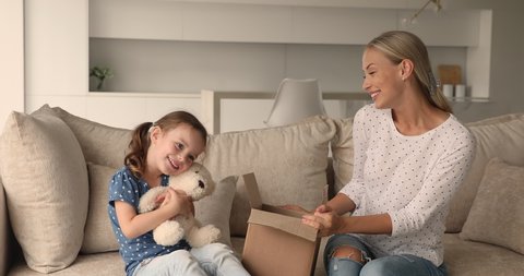Woman and daughter sit on sofa opening parcel, happy kid take out of box fluffy toy bear play with mom at home feels glad by received surprise, family use express delivery, e-commerce services concept