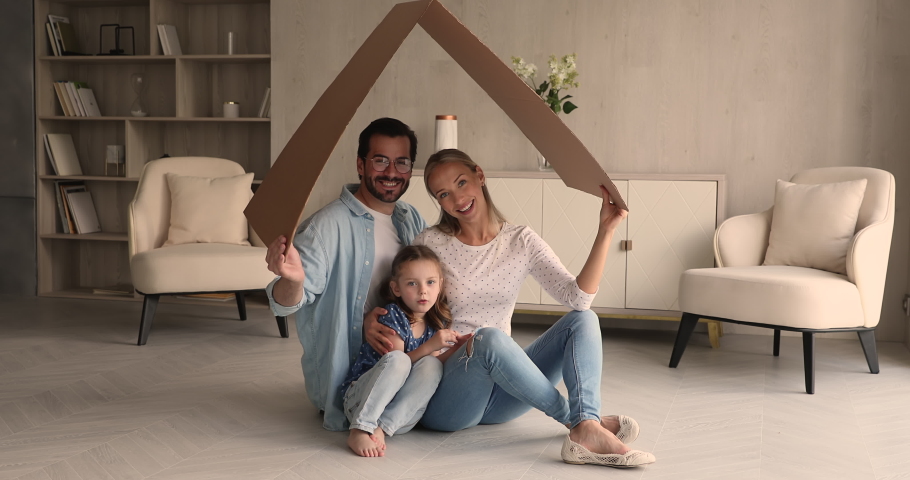 Young happy spouses their little preschooler daughter sit on floor in modern light warm living room under carton roof smiling looking at camera. New dwelling symbol, portrait family homeowners concept Royalty-Free Stock Footage #1069059451