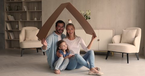 Young happy spouses their little preschooler daughter sit on floor in modern light warm living room under carton roof smiling looking at camera. New dwelling symbol, portrait family homeowners concept