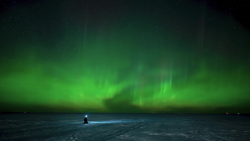 Person sitting on a frozen snow covered lake watching active green Aurora.  The subject is wearing a white head lamp.
