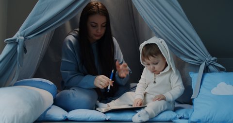 the young mother, in a tent at home with the small child, teaches her son at home to write and draw with a pen. Preschool education at home. Medium shot in slow motion.