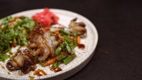 A moving video of the white ceramic plate with ginger, roasted vegetables, and octopuses pouring with soy sauce and sesame.