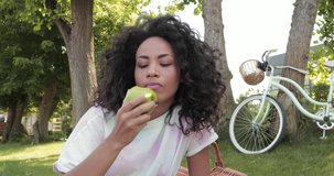 Smiling african american woman eating fresh apple during picnic in park