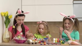 Three girls paint Easter eggs with paint and paint brushes. On the table is a golden vase with yellow tulips and a pink basket. 4k video.