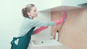 A young beautiful girl wipes the tiles in the kitchen with a rag, wearing rubber pink gloves. Cleaning the kitchen.